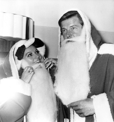 Actors Luisa Mattioli and her husband Roger Moore don "Father Christmas" costumes at the Variety Club of Great Britain's Christmas luncheon at the Savoy Hotel in London, Dec. 9, 1969. They are collecting gifts for sick and deprived children. (AP Photo/Bob Dear)
